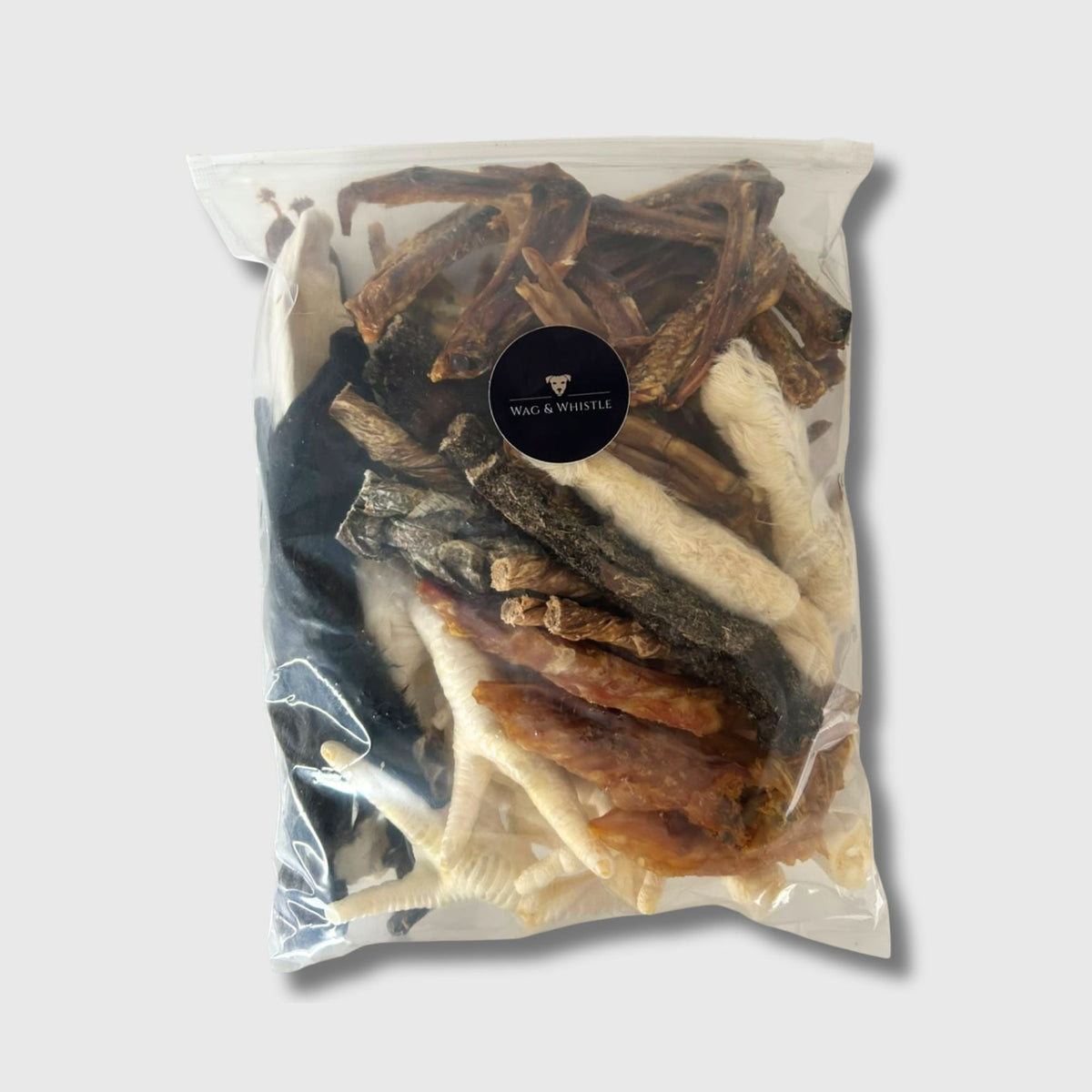 A sealed Wag & Whistle bag packed with fifty mixed natural dog treats, with chicken feet, rabbit ears and dried strips visible through the front